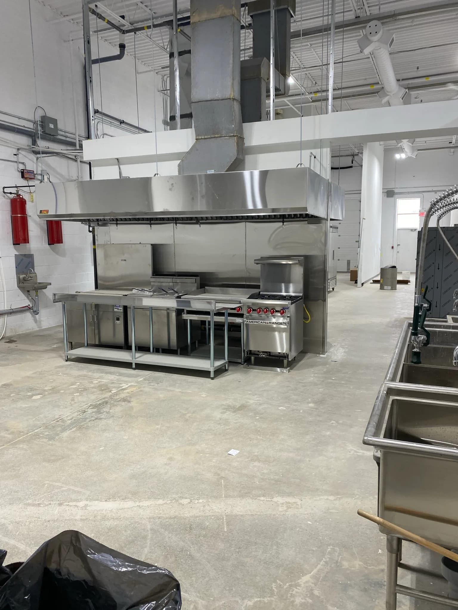 Stainless-steel commercial kitchen hood and vertical exhaust duct above cooking equipment in an installation-stage APEX project
